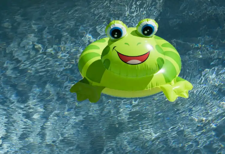 A green inflatable pool float shaped like a smiling frog is floating on clear blue water.