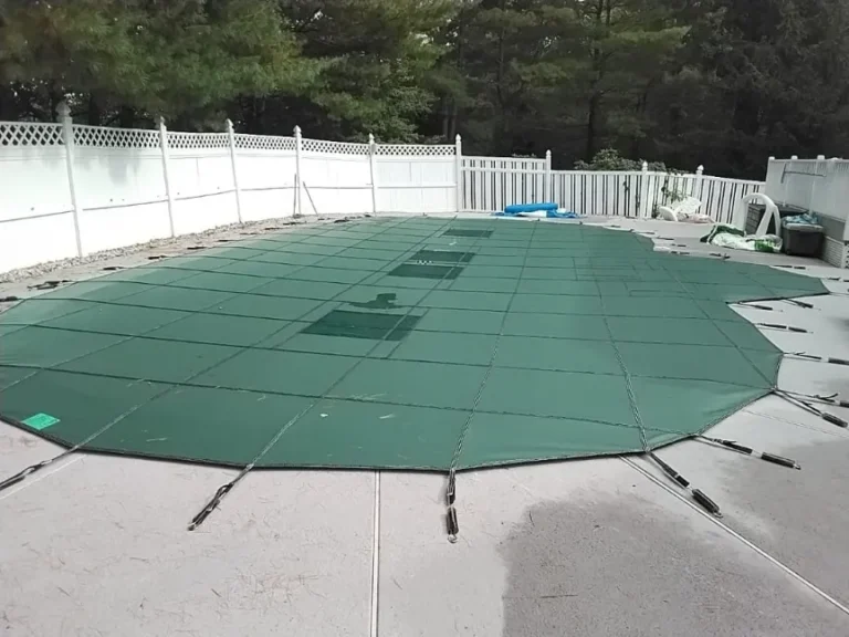 A swimming pool covered with a green safety cover secured by straps on a concrete patio, surrounded by a white fence and trees.