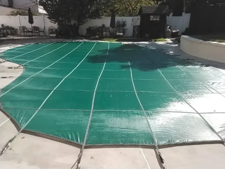 A backyard swimming pool covered with a green safety cover, surrounded by a concrete patio and outdoor furniture.