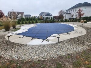 A backyard pool is covered with a blue safety cover, surrounded by rocks and landscaped plants, with houses visible in the background on a cloudy day.
